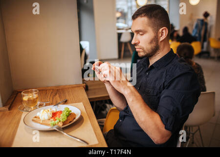 Handsome man is sitting in cafe and taking a picture of meal he ordered. He is doing that on the phone. Guy looks very serious and thoughtful Stock Photo