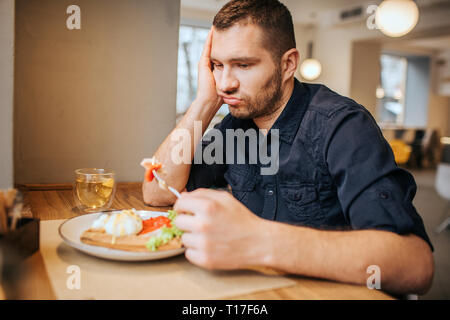 Bored and sad man is sitting at table and cafe. He is holding a piece of vegetable on fork. Man is looking at it and breathing out Stock Photo