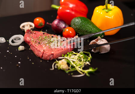 Raw steak topped with mixed herb and spice seasoning on a black background with fresh vegetables for a salad with someone reaching for the meat with t Stock Photo