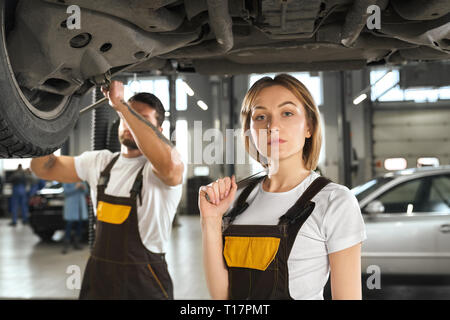 Beautiful, pretty woman wearing in white t shirt and coveralls looking at camera, posing, holding wrench. Professional mechanic fixing car undercarriage. Man and woman working in autoservice. Stock Photo