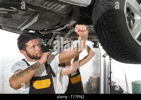 Muscular, brutal man repairing automobile with woman in autoservice. Handsome man with tattoo on hands holding tool, fixing car wheel. Mechanics working in autoservice. Stock Photo