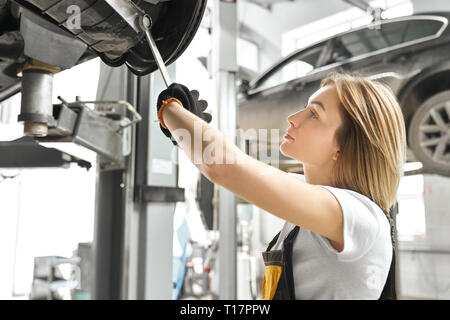 Professional female mechanic repairing automobile undercarriage, using special tool, wrench. Pretty young woman in white t shirt looking up, fixing car, working in autoservice. Stock Photo