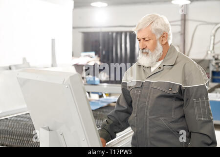 Professional engineer wearing in protective clothes, working in factory with metalwork and laser plasma cutting. Elderly bearded man operating computerized machine. Stock Photo