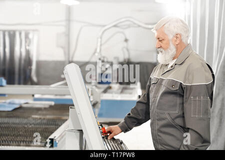Edlerly bearded man operating laser plasma cutter on metalworking factory. Experienced engineer wearing in grey uniform working with computerized machine. Stock Photo