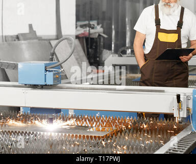 CNC laser plasma cutting metal, sparks. Engineer standing behind and observing process of plasma torch cutting steel sheet. Worker wearing in coveralls, white t shirt, holding folder. Stock Photo