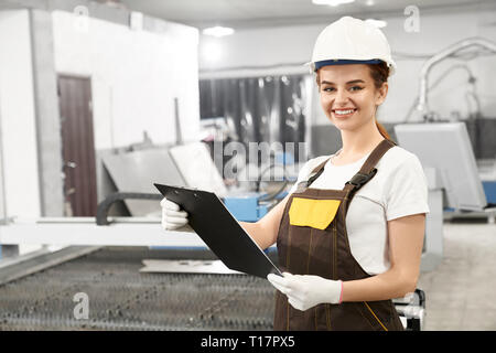 Female engineer of factory standing near plasma laser cutter. Beautiful woman wearing in white helmet, t shirt and coveralls, holding folder, looking at camera and smiling. Stock Photo