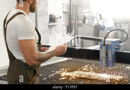 Male engineer controlling process of cutting steel. CNC plasma laser cutting metal sheet with sparks. Brutal man with tattoo wearing in coveralls and white t shirt, holding folder. Stock Photo
