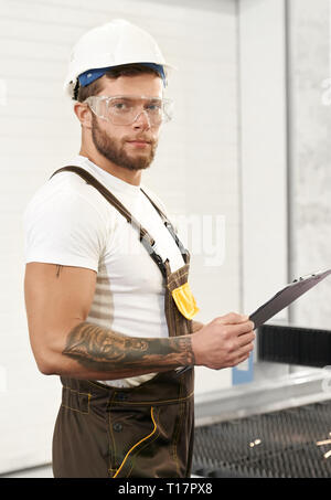 Serious, brutal man looking at camera, holding folder, posing on factory. Engineer with tattoo on hand wearing in white t shirt and hardhat, coveralls, protective safety glasses. Stock Photo