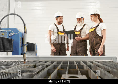 CNC plasma laser cutter on metalworking factory. Three professional workers standing, talking, looking at folder. Engineers wearing in coveralls, white helmets and t shirts. Stock Photo