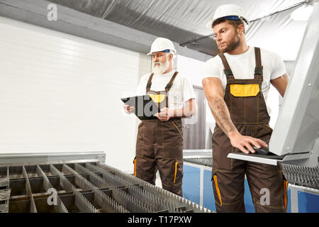 Experienced engineers of factory wearing in white helmets and t shirts, brown coveralls. Elderly man holding black folder, observing laser plasma cutter, young man operating computerized machine. Stock Photo