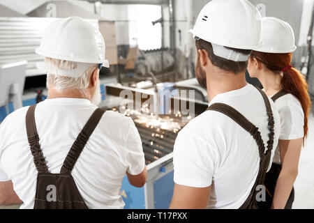 Back view of three engineers observing process of plasma laser cutting metal sheet with sparks. Workers wearing in white t shirts, white hardhats and coveralls. Stock Photo