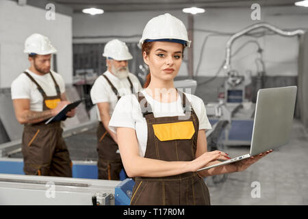 Metalworking factory. Beautiful woman worker posing, looking at camera, holding notebook. Engineers wearing in white helmets, t shirts, brown coveralls. Men working behind. Stock Photo