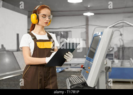 Serious, concentrated female engineer holding folder, operating computerized plasma laser machine. Beautiful woman wearing in white t shirt, gloves, coveralls, protective ear muffs and glasses. Stock Photo