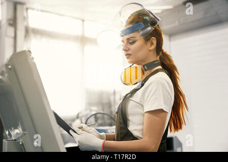 Side view of beautiful woman, engineer wearing in white t shirt, gloves, coveralls, protective ear muffs, face shield. Female worker standing near computer, holding folder. Stock Photo