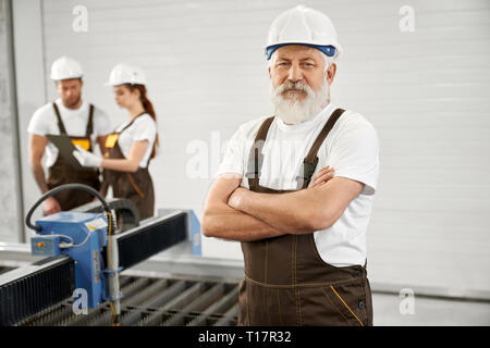 Professional elderly man posing with crossed hands on factory, Workers wearing in white helmets and t shirts, brown coveralls. Engineers standing near plasma laser cutter, talking. Stock Photo