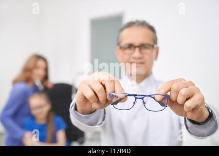 Close up of blue glasses. Doctor ophthalmologist holding, showing glasses. Medical worker in white coat looking at camera, standing in medical room of clinic. Patients on background. Stock Photo