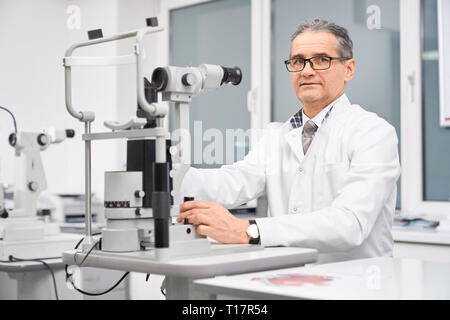 Professional doctor ophthalmologist sitting at table with special equipment slit lamp. Special device for examining eyesight of patients in medical clinic. Concept of health care and eye testing. Stock Photo