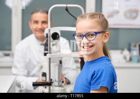 Pretty patient in blue glasses looking at camera, smiling. Cute girl testing eyeshight with slit lamp on consultation. Doctor in white coat posing, sitting in medical room of clinic.