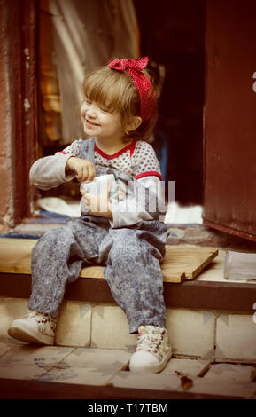White cute toddler girl wearing bright red bandana on a had and retro style denim drinks cocoa sitting on a house threshold. Happy child portrait. Stock Photo