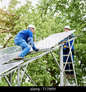 Installing of stand-alone solar photo voltaic panel system. Two workers assembling solar modules on metal platform on green tree background. Alternati Stock Photo