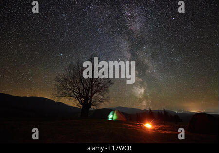 Bright bonfire burning between two tourist tents, dark and lit from inside. Camping in mountain valley under beautiful dark starry sky on distant moun Stock Photo