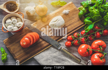 Italian food. Wooden cutting board surrounded by cooking ingredients, vegetables and mozzarella cheese on gray background. Stock Photo