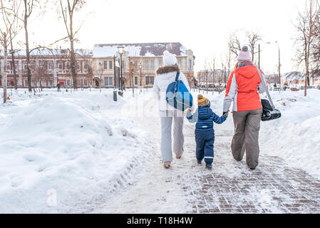 Two girlfriends little boy 3-5 years old, winter city against backdrop snow and snow drifts, view from rear, return home after training, rest on day Stock Photo