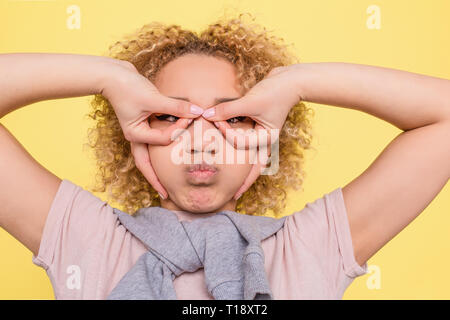 A picture of funny girl putting her fingers in a circle position and holding some air in her mouth. She looks funny but serious. Isolated on yellow ba Stock Photo