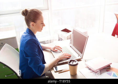 Picture of business woman in blue jacket sitting at the table near bright window and working. She is typing on keybord. Girl is wokring on the Interne Stock Photo
