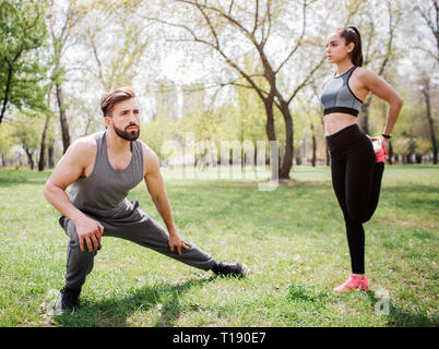 Beautiful couple is stretching outside in park. Guy is squating and stretching his legs while his girlfriend is standing and stretching her legs Stock Photo
