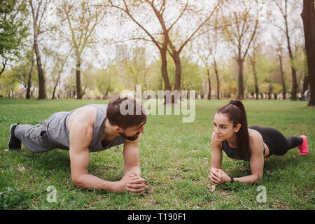 Good partners are standing in a plank position on grass. Man and woman are looking to each other and smiling. This couple is very cheerful Stock Photo