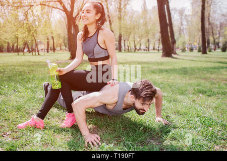 Funny picture of young man and woman. He is doing push ups and looking straight while she is sitting at him and laughing. Also girl is holding a bottl Stock Photo