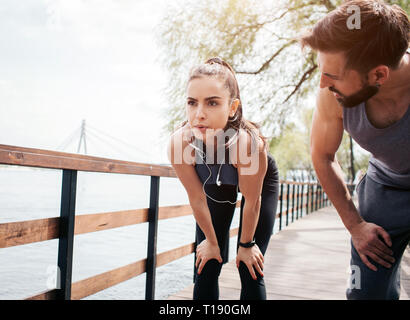 Attractive girl is tired of jogging. She has lost her breath. Girl is having some rest. Guy is stnading besides the girl and looking at her Stock Photo