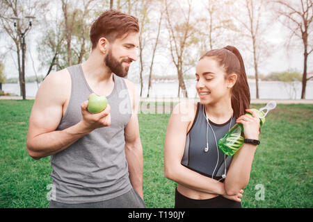 Young and healthy couple are standing and looking to each other and smiling. He is eating an apple. Girl has a headphones around her neck and holding  Stock Photo