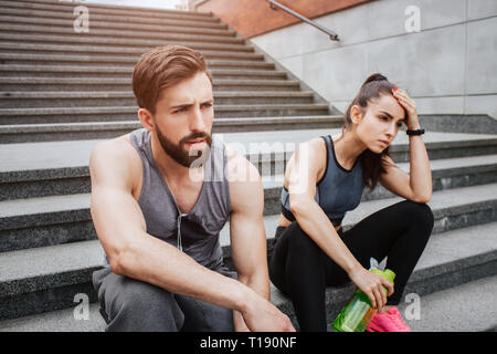 Very tired couple is sitting on steps and having some rest. They have run a lot. Girl is keeping her hand close to the head while her boyfriend is jus Stock Photo