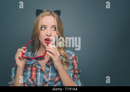 Portrait of a girl wears kitten ears. Also she is holding dark glasses with red edge and looking to the right. She is biting a small piece of glasses. Stock Photo