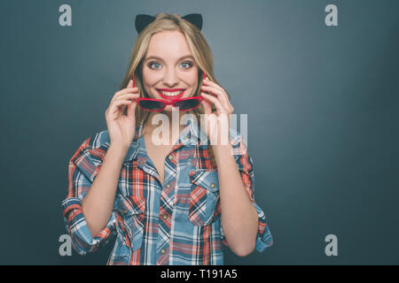 Nice and happy person is wearing grey with red shirt and a kitten ear on her head. Also she is putting on her glasses and smiling. Girl is looking str Stock Photo