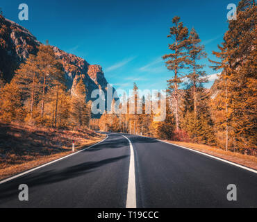 Road in autumn forest at sunset in Italy. Beautiful mountain roadway, orange tress, grass, high rocks, blue sky. Landscape with empty asphalt road thr Stock Photo