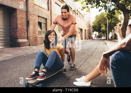 Woman sitting on skateboard being pushed by a male friends while other friends sitting by talking their pictures with smart phone. Friends having fun  Stock Photo