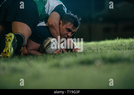 Rugby player being blocked by opposite team players. Sports men pins down opponent during rugby match at night. Stock Photo