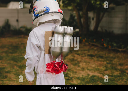 Rear view of boy in space helmet and suit carrying a toy jetpack on his back. Boy pretending to be an astronaut playing outdoors. Stock Photo