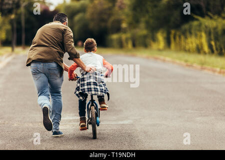 Rear view of a boy riding a bicycle while his father runs along holding the kid. Father teaching his son to ride a bicycle. Stock Photo
