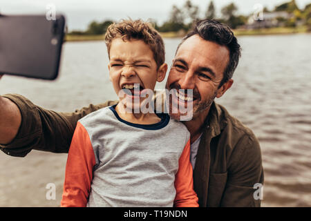 Close up of father and son taking a selfie near a lake. Happy kid making faces sticking his tongue out while his father takes a selfie. Stock Photo