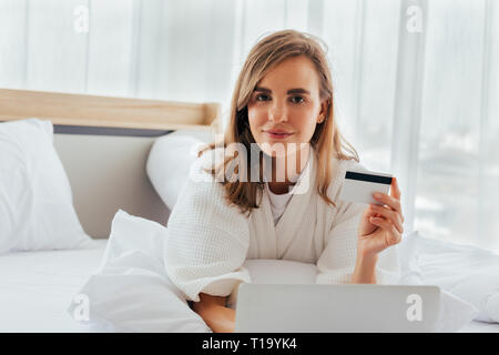 Young positive woman showing plastic card near laptop and lying on bed in light room Stock Photo
