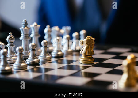 Golden knight placed against many silver chess pieces on chessboard with businessman in background Stock Photo