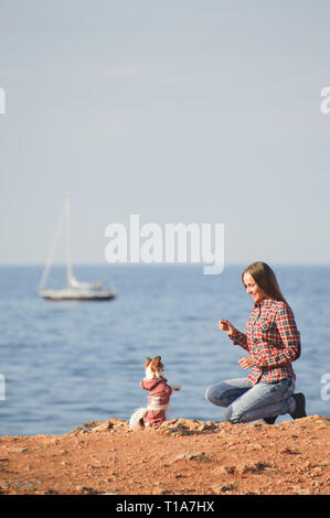 happy young girl in plaid shirt and jeans training chihuahua little dog to sit on sea coast with sailing boat during outdoor leisure Stock Photo
