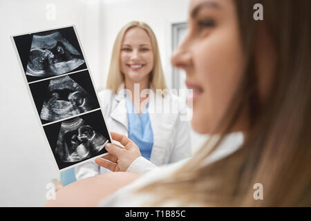 Happy future mother holding x ray, looking away. Cheerful, professional doctor smiling. Consultation of pregnant woman and therapist in medical clinic. Stock Photo