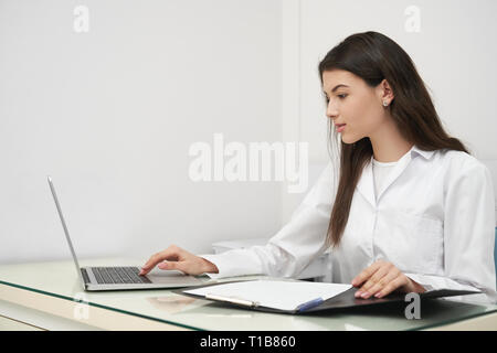 Beautiful woman, doctor sitting at table and working, using notebook and folder. Young medical specialist posing in medical office. Concept of medicine, hospital and private clinic. Stock Photo