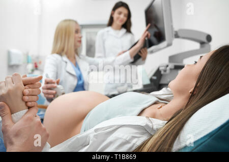 Pregnant woman lying on sonography diagnosis in clinic. Female doctor and nurse doing ultrasound examination of woman's abdomen, pointing with finger at monitor of modern machine. Stock Photo