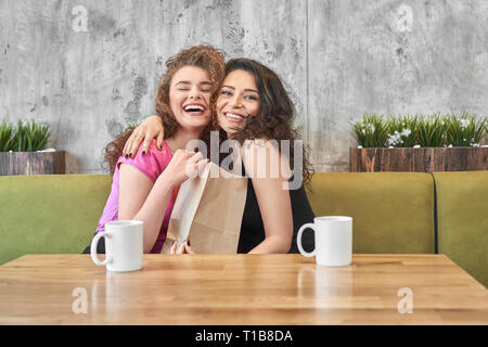 Happy pretty girl hugging friend. Cheerful young women spending time together in cafe, hanging out, sitting on sofa at table with white mugs. Female friends smiling, holding paper bag with present. Stock Photo